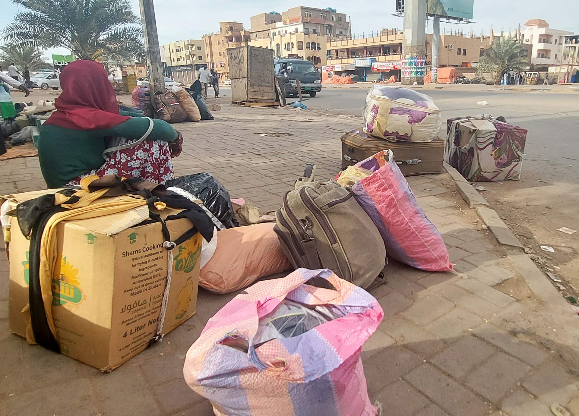 Eine Frau sitzt auf einem Karton auf einer Straße. Um sie herum stehen Taschen.