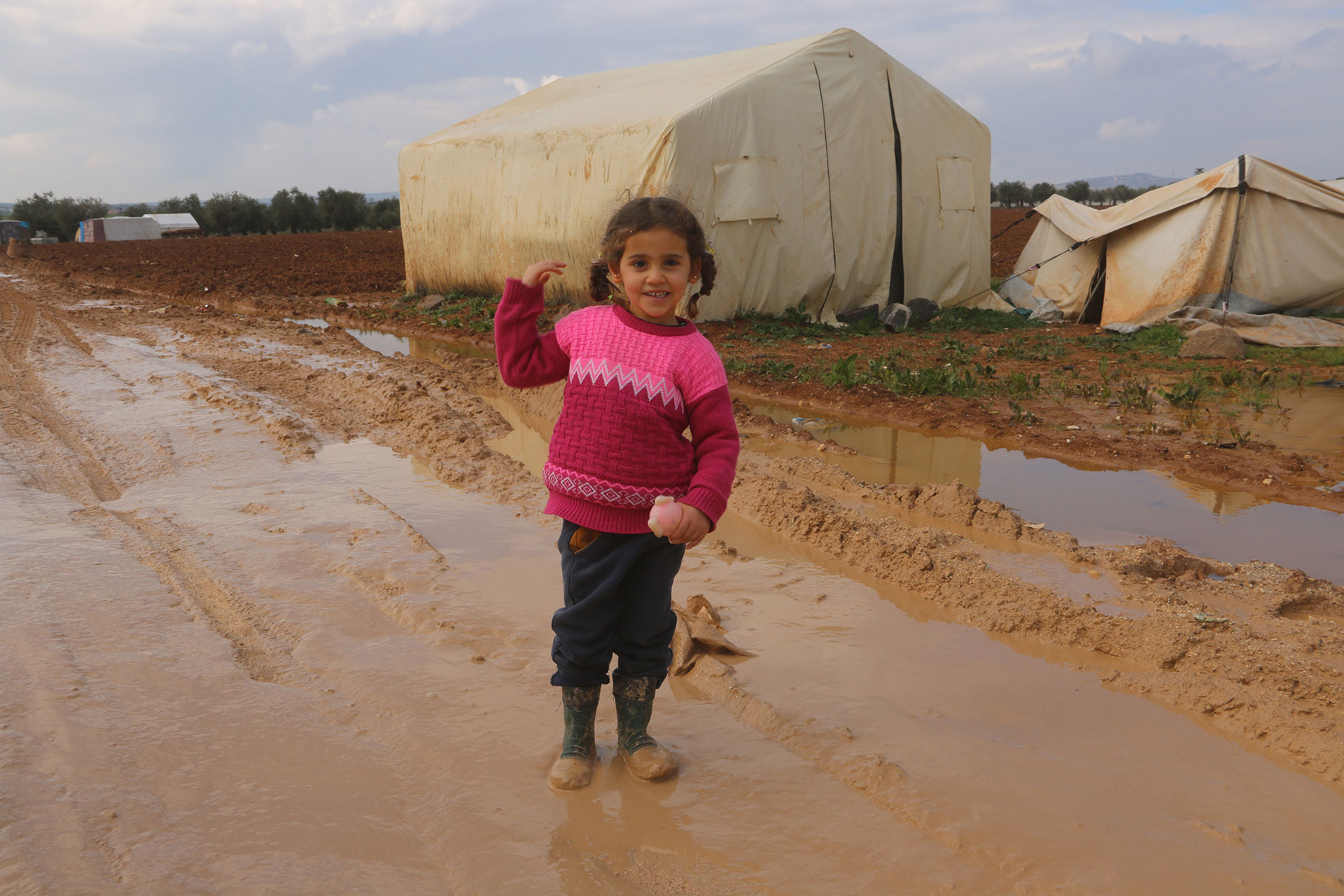 Ein lachendes Mädchen in Gummistiefeln steht auf einer matschigen Straße. Im Hintergrund sieht man ein weißes Zelt.