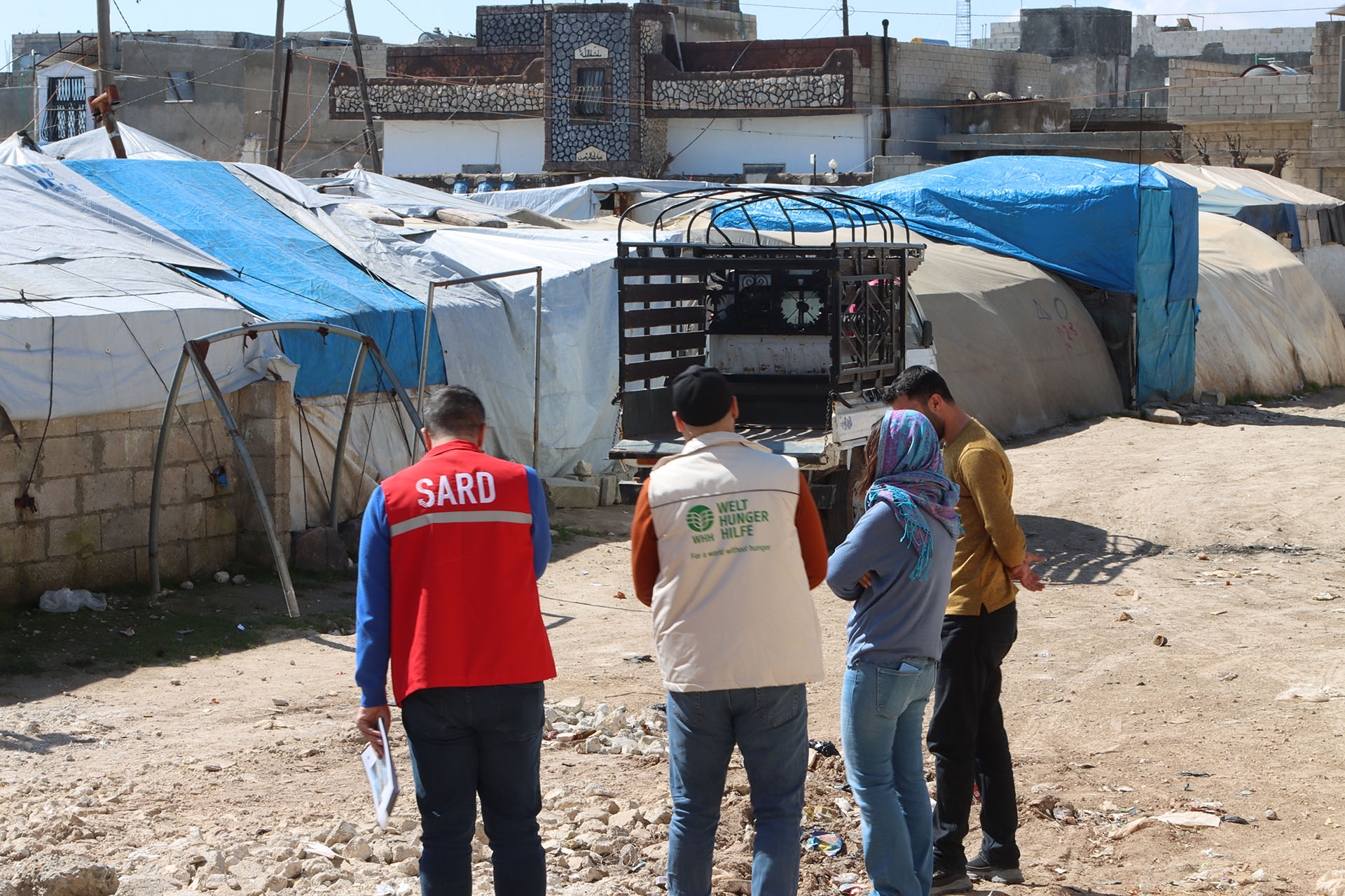 Vier Personen, zwei von ihnen in Uniformen von Hilfsorganisationen, in einem Flüchtlingslager in Syrien. Die Umgebung besteht aus provisorischen Unterkünften, die mit blauen und weißen Planen bedeckt sind.