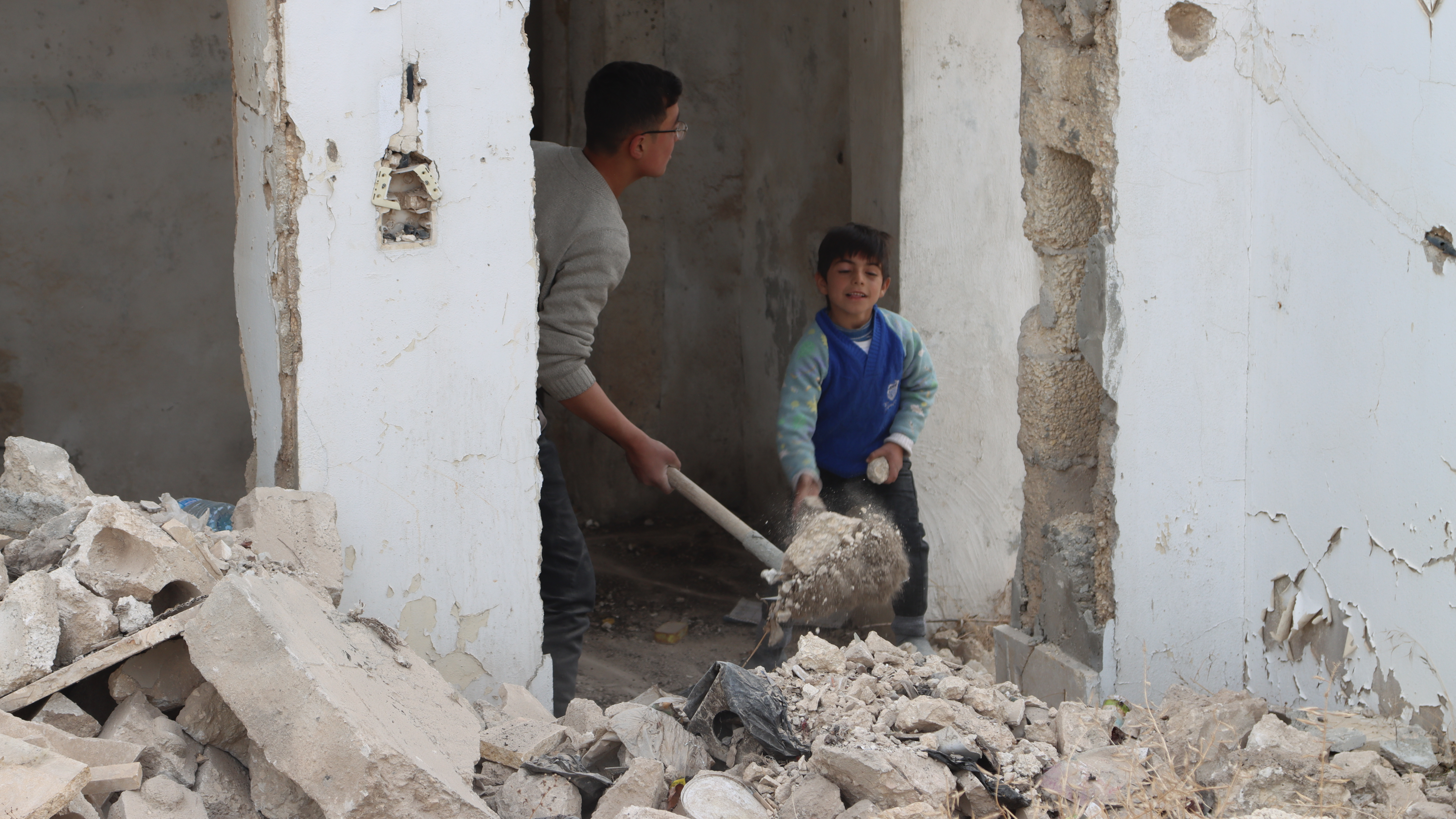 Ein älterer und ein jüngerer Junge räumen mit Schaufeln Schutt aus einem zerstörten Haus.