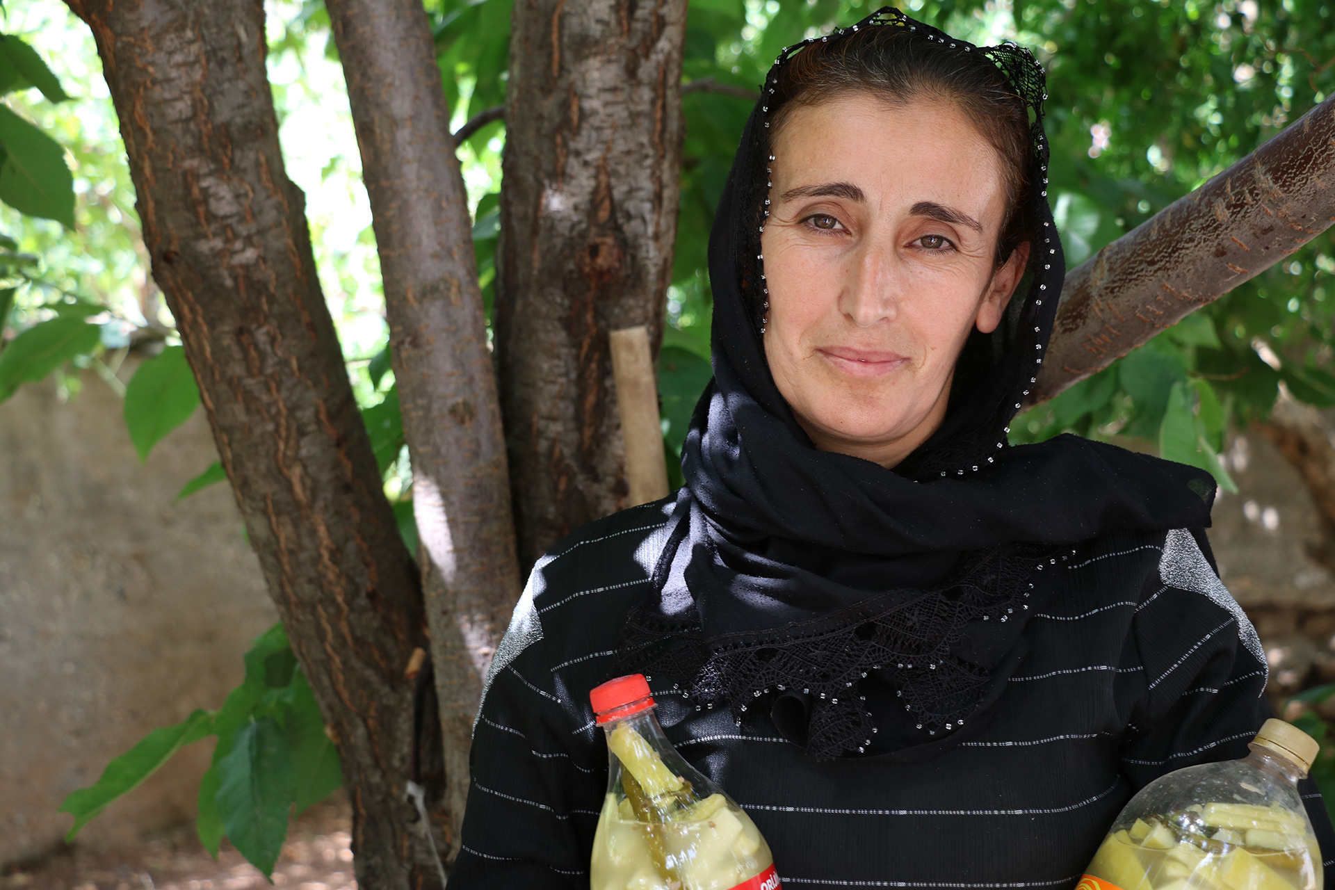 Eine Frau steht unter einem Baum, in der Hand hält sie zwei Flaschen mit Gewürzgurken, Türkei 2021.