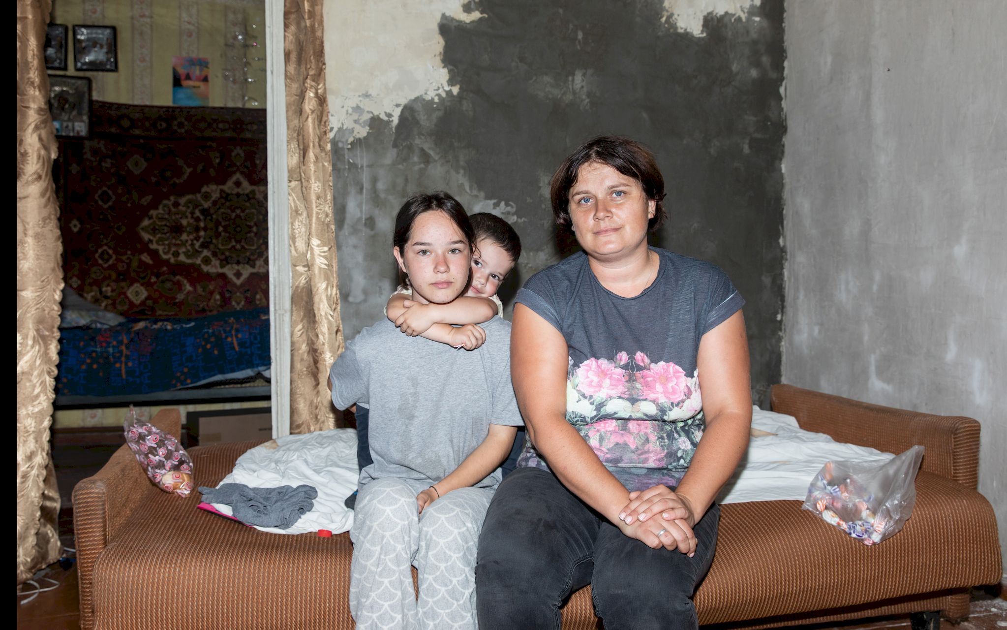 In einer kargen Wohnung sitzen eine Frau und zwei Kinder auf der Kante des Bettes.