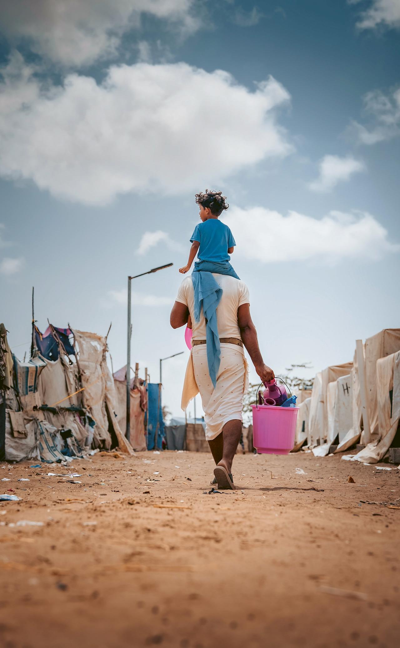 Ein Vater trägt sein Kind auf den Schultern und hält einen Eimer mit verschiedenen Gegenständen in der Hand.